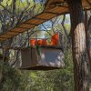 Una tenda sospesa con sedie arancioni sopra, fissata tra gli alberi con passerelle di legno e reti.