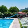 Piscina con scivolo azzurro, sdraio bianche su prato verde e ombrellone rosso.