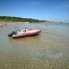 Gommone rosso-nero nelle acque limpide, spiaggia e bagnanti sullo sfondo.