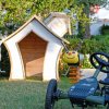 Un kart giocattolo su prato, vicino a giochi a forma di lumaca e ape e una casetta di legno.