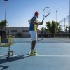 Giocatore di tennis colpisce la palla sul campo. Un carrello con racchette è vicino.